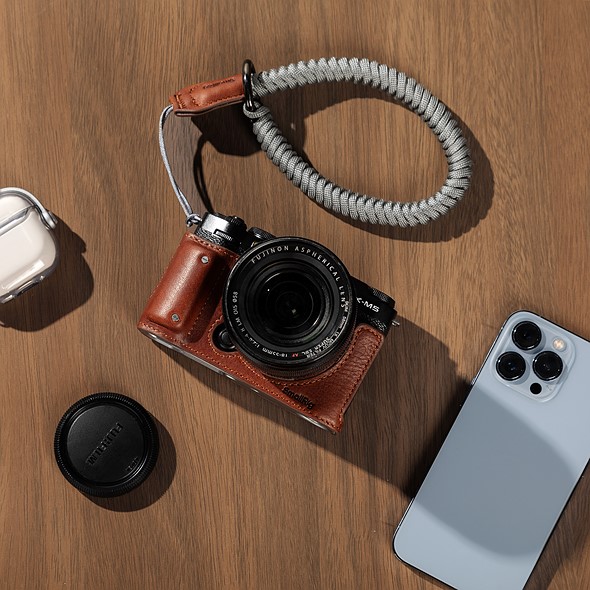 Photo of a Fujifilm X-M5 in a brown half-leather case laying on a wood table with a body cap, iPhone, and Airpods