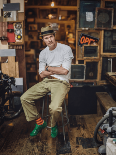 A stylish man in a hat and colorful socks sitting on a stool in a rustic workshop, photographed with the Brenizer method for a wide, shallow-depth portrait with creamy background blur.