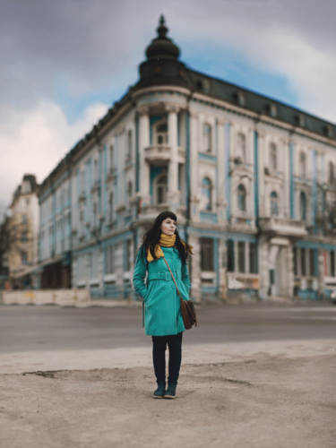 A woman in a teal coat and yellow scarf stands confidently in front of a historic European-style building, with the Brenizer method used to isolate the subject against the urban backdrop.