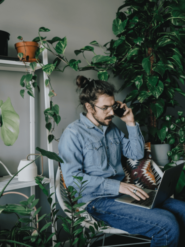 A man with a bun and glasses works on a laptop and talks on the phone while seated in a plant-filled room, representing environmental portraits that blend personal style and workspace.