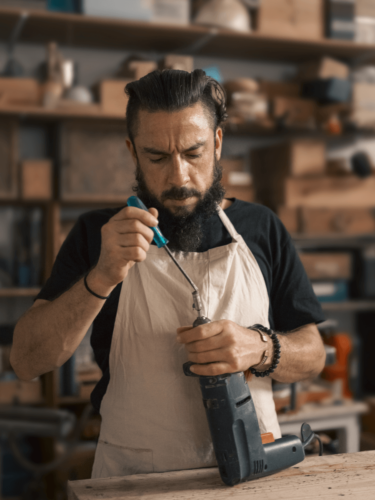 A focused man in an apron uses a screwdriver to repair a power drill in a cozy, tool-filled workshop.