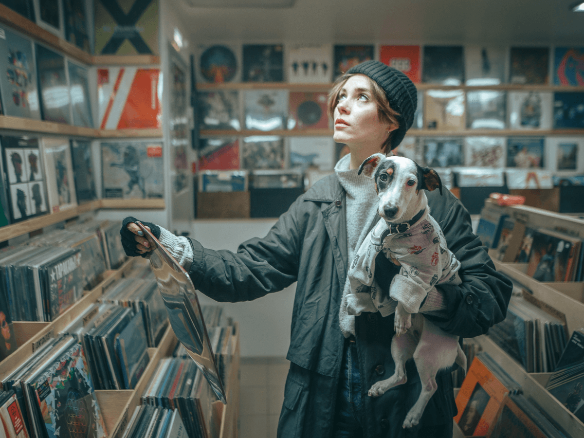 A woman in a knit beanie holds her dog while browsing vinyl records in a colorful, retro record store.