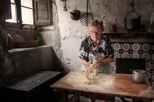 elderly woman shakes pasta in rustic kitchen