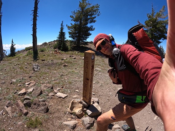 person stands next to trail marker in sparse forest with blue skies