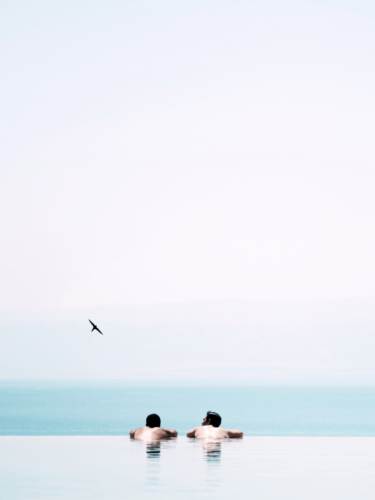 Two people relax in an infinity pool overlooking a calm sea and sky, reflecting simplified photography with serene composition and negative space.