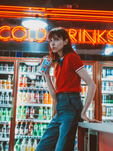 A young woman in a red shirt sipping an ICEE drink inside a neon-lit convenience store, styled with film emulation to replicate a retro, cinematic look.