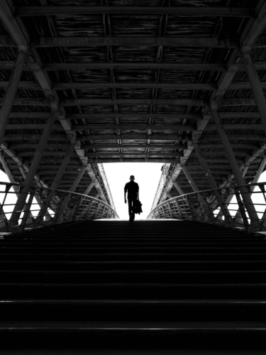 A lone figure ascends a dark staircase under a bridge, evoking a sense of dramatic storytelling.