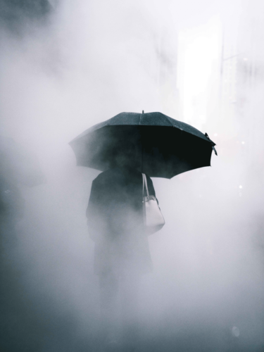 A person holding an umbrella walks through a thick, foggy city street, creating a mysterious and moody foggy scene.