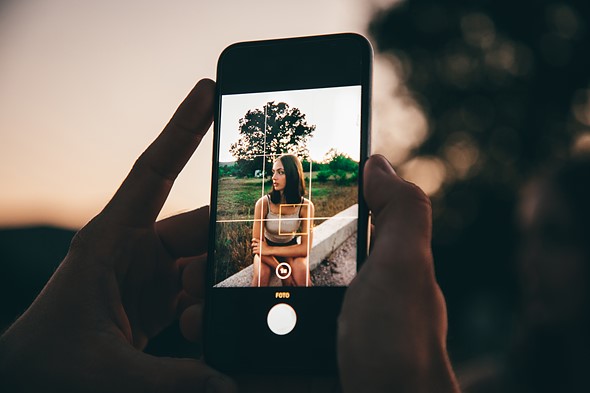 hands hold a phone while taking a photo of a woman in front of a field and tree