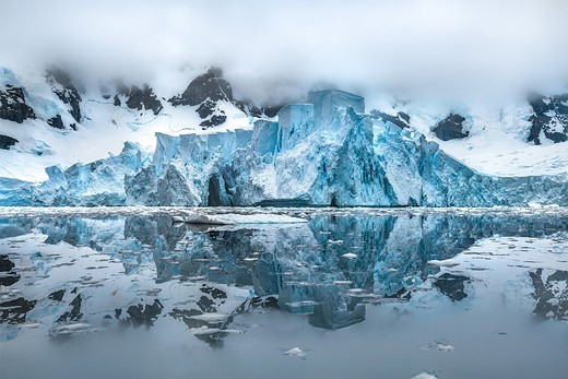 icy cliffs shrouded in clouds loom over iceburg filled water
