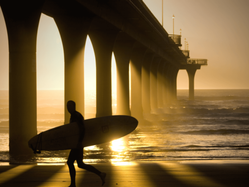 A surfer silhouetted against a golden sunset, walking beneath a pier with long shadows on the beach.