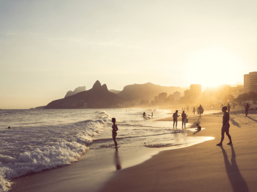 Silhouettes of people walking along a beach at sunset with waves rolling in, captured with nostalgic color grading that softens the scene with warm, dreamy light.