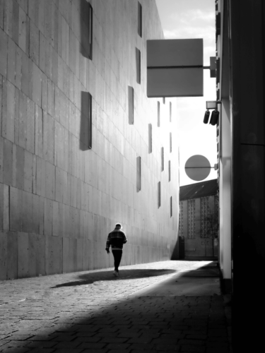 Dramatic contrast in black-and-white photography capturing a lone man walking down a sunlit alleyway, emphasizing shadows and light.