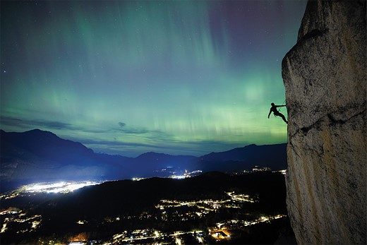a climber hangs from a cliff overlooking city lights with northern lights in the sky