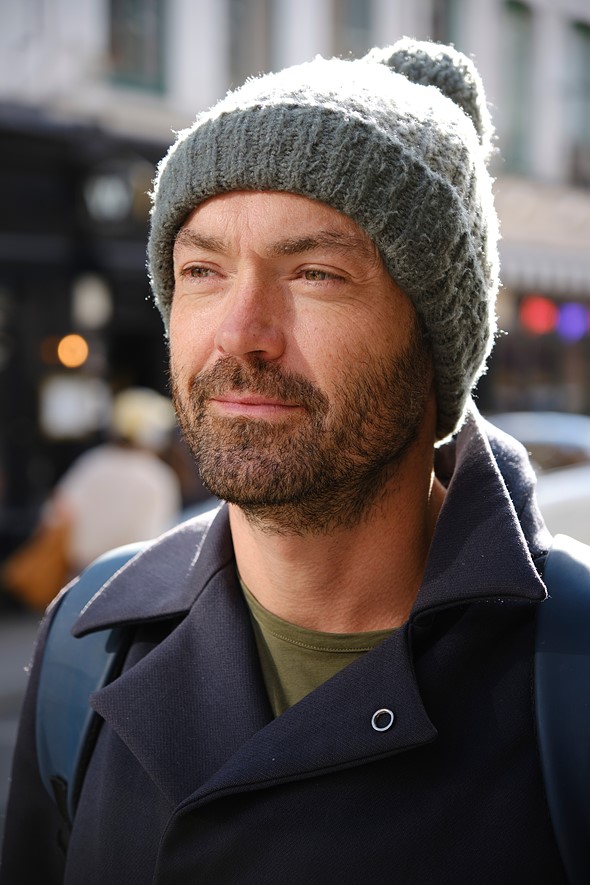 a man with a gray winter hat gazes into the distance on a sidewalk in a city