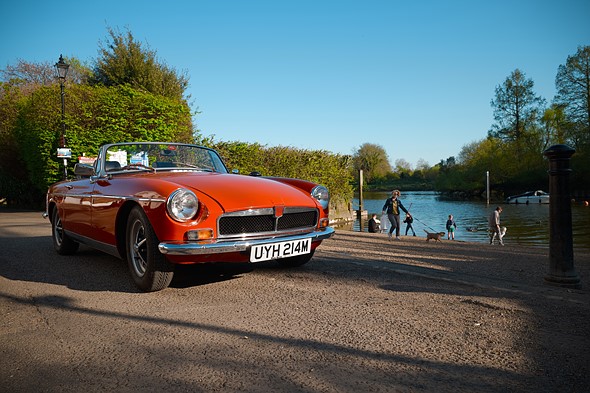 a vintage red convertible is parked next to a waterfront park with people walking