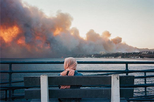 a woman sits on a bench with smoke and fire in the distance