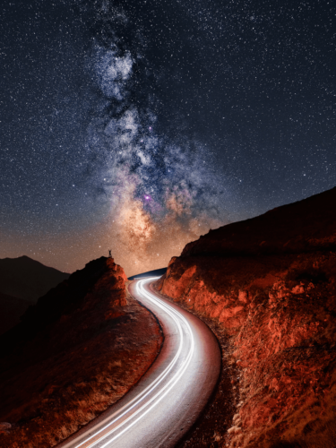 A winding mountain road leading to the Milky Way, with car light trails captured through long exposure photography to enhance the night sky scene.