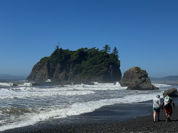 people walk on a black sand beach with large rocks in the water