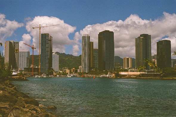 skyscrapers  and cranes sit beyond a bay with boats with green mountains in the background