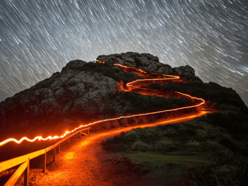 A glowing path up a mountain under star trails, captured with long exposure to convey expressive motion through swirling sky and light trails.