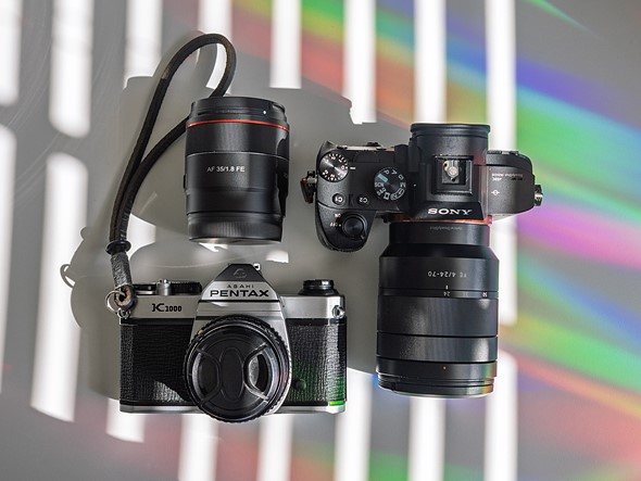 two cameras and a lens sit on a white desk with rainbow light and shadows from blinds