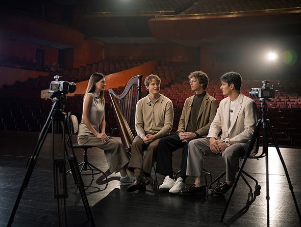 a group of four people sits on a stage in a theater with two cameras on tripods on either side