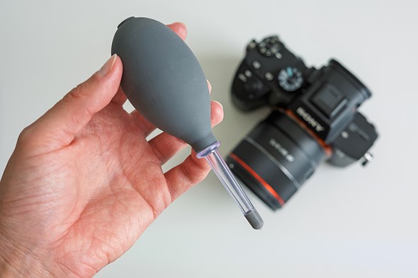 a hand holds a bulb blower over a camera on a white desk