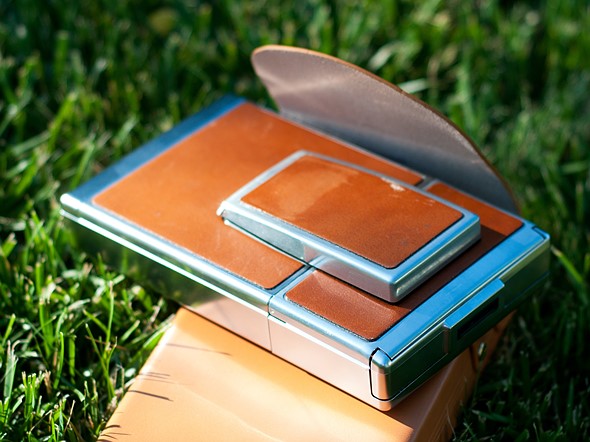 a leather and metal camera is folded into a rectangle while placed on a leather case on grass