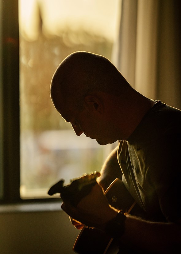 a man sits with their head looking down while playing a guitar in front of a window with golden light streaming in