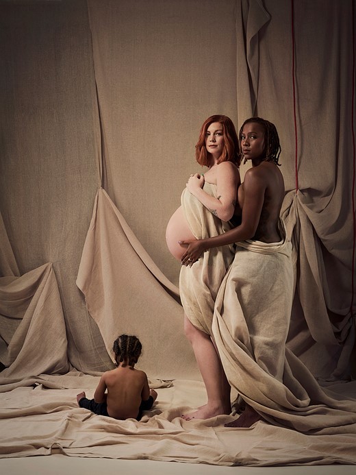 a young child sits on the ground next to two women embracing in front of curtains