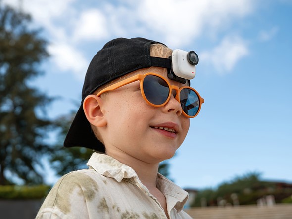 a young child with orange sunglasses smiles while wearing a hblack hat with small action camera attached