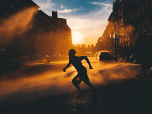 A boy dashing through a street sprinkler at sunset, with golden light creating contrast and depth, characteristic of golden hour portraits.