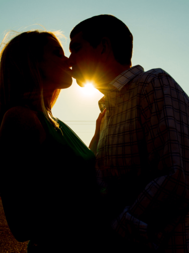 A couple kissing with the setting sun perfectly aligned between them, creating a dramatic burst of light that emphasizes sun flare.