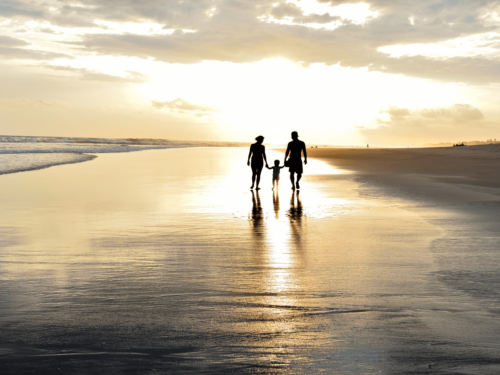 A family holding hands while walking along a reflective beach at sunset, creating a soft glow effect that defines artistic backlight.