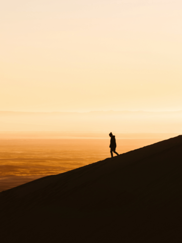 A lone figure walking along a desert dune at sunset, with warm tones highlighting the scene, representing summer backlighting photography.