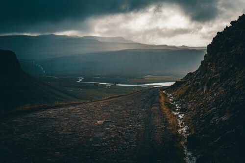 Moody mountain road landscape by Nicole Grenier, with a rugged dirt path leading into misty mountains under dramatic storm clouds.