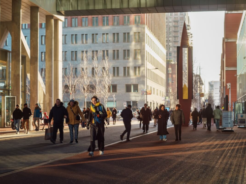 People walking through a modern city street in The Hague during golden hour, with warm sunlight casting long shadows between tall buildings, captured by Sirun Tang.