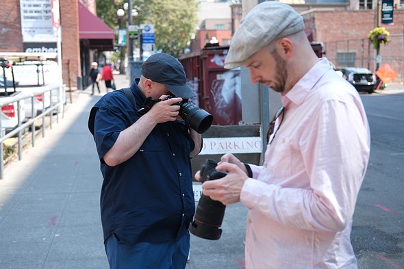 two men stand looking at cameras on a sidewalk