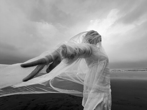 Winning photo from the 2025 OnePlus Photography Awards showing a person on a beach draped in translucent fabric, captured in black and white.