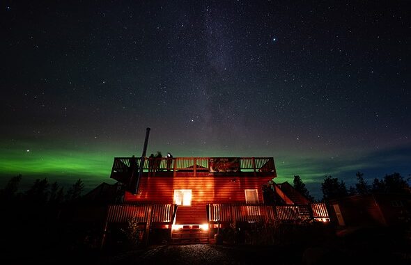 a_log_cabin_is_lit_up_and_glowing_orange_beneath_a_night_sky_filled_with_stars_and_aurora