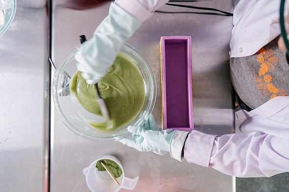 a person wearing light blue rubber gloves stirs a bowl with green liquid in it on a stainless steel table with purple mold