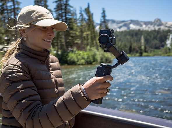 a woman wearing a brown puffy jacket stands holding a gopro gimbal on a bridge over a mountain river