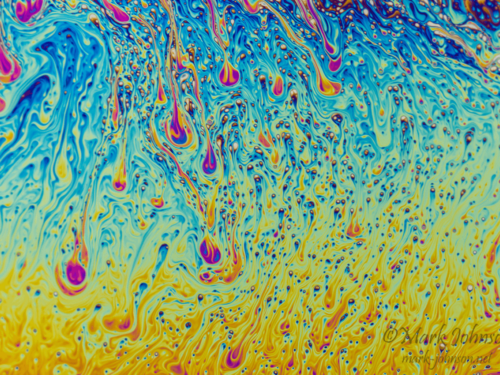 Abstract macro image of swirling colorful liquid patterns in blue, yellow, and pink, resembling a soap film surface.