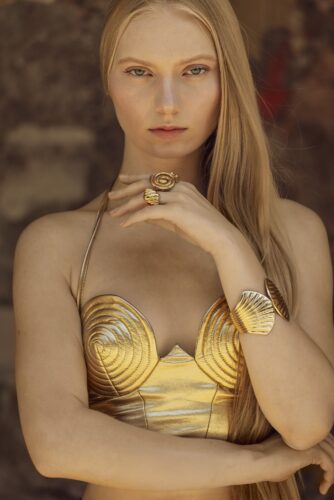 Fashion portrait by Olena Leliuk of a woman in a sculptural gold bodice with matching jewelry, her long blonde hair flowing over one shoulder.