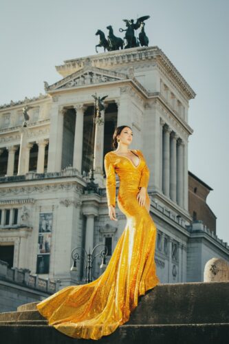 Fashion portrait by Olena Leliuk of a woman in a dramatic golden gown posing in front of a neoclassical building with statues in Rome.