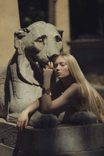 Artistic portrait by Olena Leliuk of a woman with long blonde hair leaning against a stone lion statue, blending elegance with ancient symbolism.
