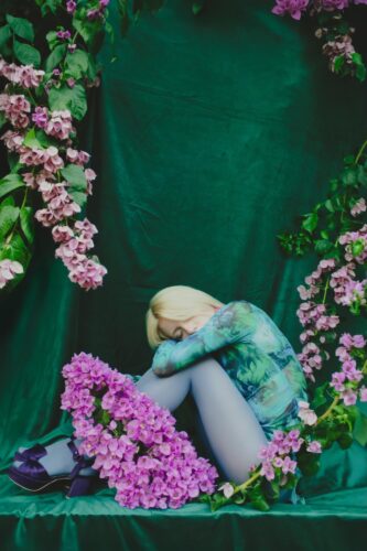 Artistic portrait by Olena Leliuk of a woman in floral-themed clothing sitting curled up against a green backdrop, surrounded by pink blossoms.