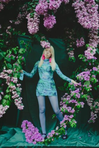 Fashion portrait by Olena Leliuk of a woman in a patterned green-blue dress and lilac tights standing amid cascading pink flowers against a dark green backdrop.