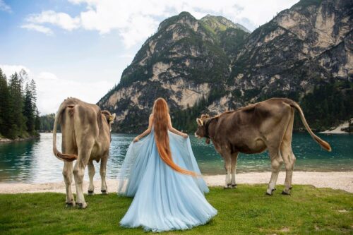 Fantasy-inspired photo by Olena Leliuk showing a woman in a flowing blue gown with long red hair standing between two cows by a mountain lake.